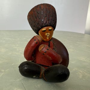 May include: A hand-carved wooden figurine of a seated person. The figure wears a dark brown hat, a red shirt, and black pants. The face is painted with tan and brown tones. The arms are red with black dots. The figurine is a folk art piece.