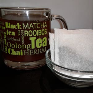 May include: A glass mug filled with dark tea, with the words "Black Tea, Matcha, Rooibos, Tea, Oolong, Chai Herbal" printed on the side. A white tea bag sits in a clear glass bowl.