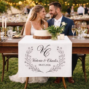 Peut inclure: Chemin de table blanc avec une couronne florale et les noms "Victoria & Charles" imprimés en noir. Un gâteau de mariage repose sur une base en bois. Un couple se sourit en arrière-plan.