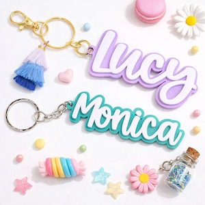 May include: Two personalized name keychains, one lavender and one teal, with the names "Lucy" and "Monica" in white script. The keychains have gold and silver hardware, with a blue and lavender tassel. Decorative elements include a pink heart, candy, and flowers.