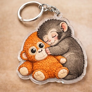 May include: A clear acrylic keyring with a cartoon illustration of two monkeys embracing. One monkey is orange, the other is grey. The keyring has a silver metal ring and chain.
