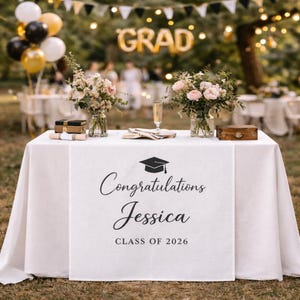 Peut inclure: Une nappe blanche avec un chemin de table sur le thème de la remise de diplômes. Le chemin de table porte le texte "Félicitations Jessica CLASS OF 2026" et un dessin de toque de diplômé. La table est décorée de fleurs, de champagne et de petites boîtes en bois. Des ballons et une bannière "GRAD" sont en arrière-plan.