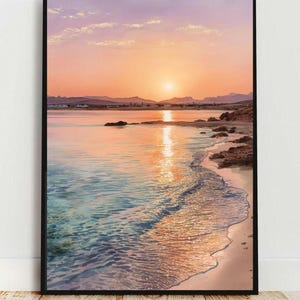 Puede incluir: Impresión enmarcada de una escena de playa al atardecer. La obra de arte representa una vibrante puesta de sol con tonos naranjas y rosas que se reflejan en el agua. La costa presenta arena y rocas, con montañas en la distancia. El marco es negro.