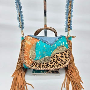 Op de afbeelding: Een kleurrijke crossbodytas met een mix van texturen en patronen. De tas heeft een turquoise, oranje en luipaardprint ontwerp, versierd met goudkleurige vleugelaccenten en lange bruine franjes. De riem is van denim en luipaardprint.