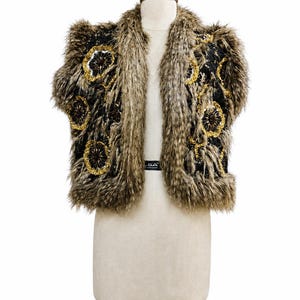May include: A sleeveless vest featuring a black base with long, shaggy brown and tan faux fur trim along the edges. The front panels are decorated with circular floral-like patterns made of gold sequins and dark brown beads throughout the fabric.