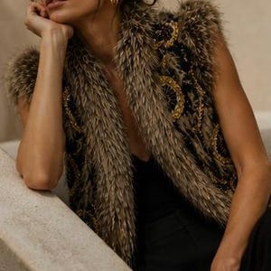 May include: A sleeveless vest featuring a dark base with intricate gold circular embroidery and a thick, textured brown faux fur trim along the collar and armholes, worn over a simple black top and black trousers with gold hoop earrings.