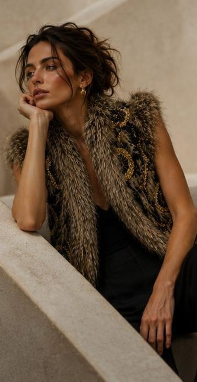 May include: A sleeveless vest featuring a dark base with intricate gold circular embroidery and a thick, textured brown faux fur trim along the collar and armholes, worn over a simple black top and black trousers with gold hoop earrings.