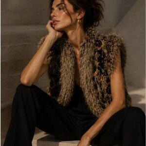May include: A sleeveless vest featuring a textured, shaggy brown and black faux fur exterior with gold-toned circular embellishments. The garment is worn over a simple black top and paired with black trousers, creating a layered appearance.