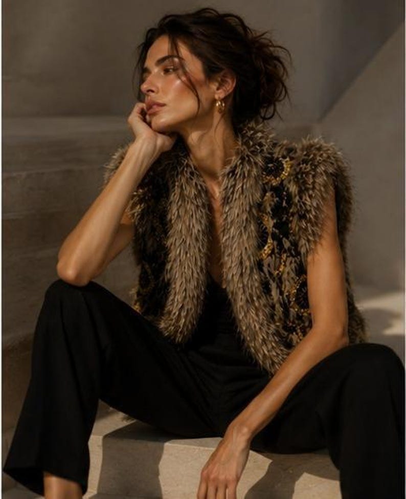 May include: A sleeveless vest featuring a textured, shaggy brown and black faux fur exterior with gold-toned circular embellishments. The garment is worn over a simple black top and paired with black trousers, creating a layered appearance.