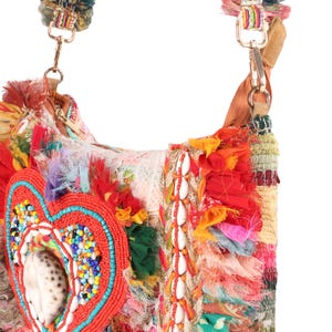 May include: A colorful, handcrafted shoulder bag with a heart-shaped embellishment. The bag features a mix of textures, including fabric scraps, beads, and seashells. The strap is adorned with tassels and gold-colored hardware. The bag's design incorporates red, orange, blue, and white.