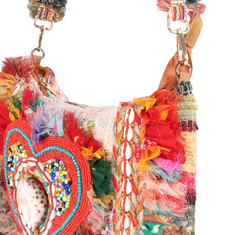May include: A colorful, handcrafted shoulder bag with a heart-shaped embellishment. The bag features a mix of textures, including fabric scraps, beads, and seashells. The strap is adorned with tassels and gold-colored hardware. The bag's design incorporates red, orange, blue, and white.
