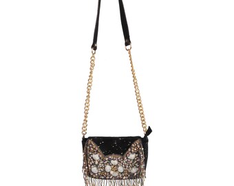 May include: Black crossbody bag with a gold chain strap. The bag features an embellished flap with white and gold floral designs and beaded fringe. The bag is made of a black fabric with a speckled pattern.