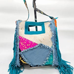 Op de afbeelding: Een patchwork crossbodytas met een turquoise franje en riem. De tas heeft een mix van materialen, waaronder denim, koeienhuid en kleurrijke accenten. Het ontwerp omvat een turquoise vierkante uitsparing en een mix van roze, blauwe en gouden patronen.