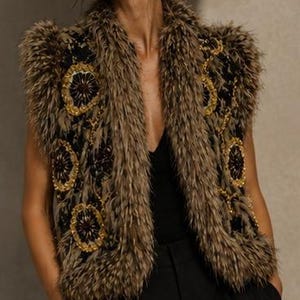 May include: A sleeveless vest featuring a textured brown faux fur trim along the edges and collar. The black base fabric is decorated with circular gold and dark brown embroidered floral patterns. The garment is worn over a simple black top.