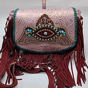 May include: A pink crossbody bag with a decorative flap featuring an eye design with turquoise and pearl accents. The bag has a burgundy fringe and strap, and gold-colored hardware. The flap has a floral embossed pattern.