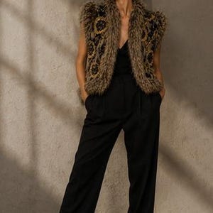 May include: A sleeveless vest featuring a textured, shaggy brown faux fur finish with dark circular embroidered patterns, worn over a black V-neck top and black wide-leg trousers, paired with black pointed-toe heels.