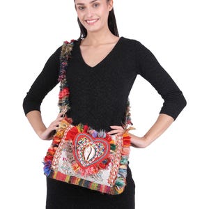 May include: A colorful, square shoulder bag with a heart-shaped design. The bag features a red heart with a seashell in the center, surrounded by beads. The bag has a white lace background and is trimmed with colorful fringe and a woven strap.