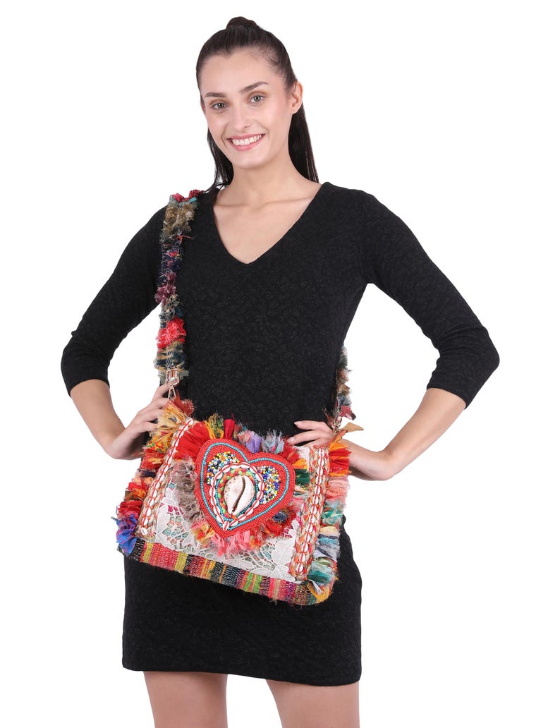 May include: A colorful, square shoulder bag with a heart-shaped design. The bag features a red heart with a seashell in the center, surrounded by beads. The bag has a white lace background and is trimmed with colorful fringe and a woven strap.