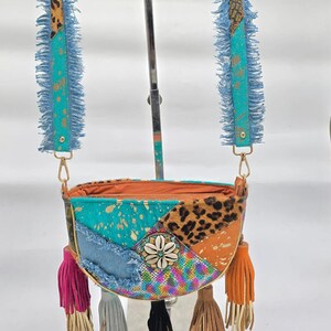 Puede incluir: Bolso bandolera patchwork con una mezcla de texturas y colores. El bolso presenta un estampado de leopardo, detalles en denim y turquesa. Tiene un adorno floral y borlas coloridas. Las correas son azules con flecos.