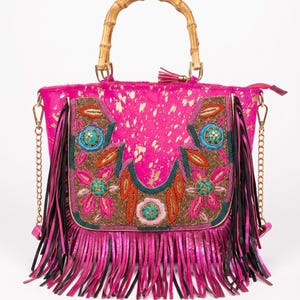 May include: A bright pink handbag with a bamboo handle and long black and pink fringe. The front features a decorative panel with intricate beadwork in floral and leaf patterns, gold metallic accents, and a detachable gold chain shoulder strap.