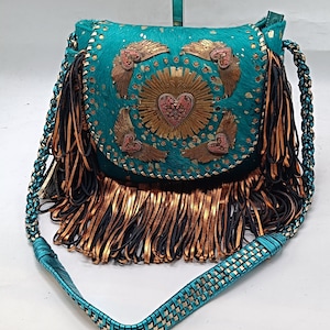 May include: A turquoise leather crossbody bag with a braided strap. The bag features a flap with a heart and wing design, and a fringe of brown and black leather. The bag is decorated with gold accents.