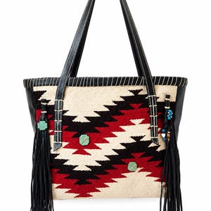 May include: A tote bag featuring a woven cream, red, and black geometric pattern. The bag has black leather straps, black leather side panels, and long black fringe tassels on both sides. Three square turquoise accents are attached to the front.