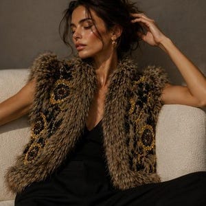 May include: A sleeveless vest featuring a shaggy brown faux fur texture, accented with black panels and intricate gold-toned circular embroidery. The garment is worn over a simple black top, showcasing a structured open-front design with a high collar.