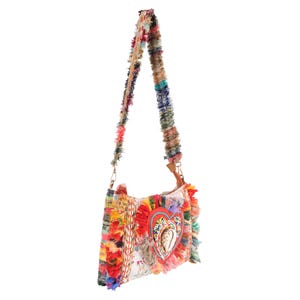 May include: A colorful shoulder bag with a fringed design. The bag features a heart-shaped embellishment with beadwork and a seashell. The strap and body of the bag are adorned with various colors of fabric. The bag is a unique accessory.