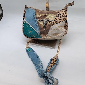 May include: A patchwork crossbody bag with a gold chain strap. The bag features panels of denim, leopard print, and metallic gold fabric. A silver bull head adorns the front. The strap is denim with leopard print accents.