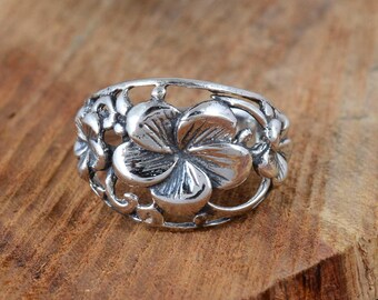 May include: A silver-colored ring featuring a floral design. The ring has a series of flowers with detailed petals and a filigree band. The ring is set against a wooden surface.