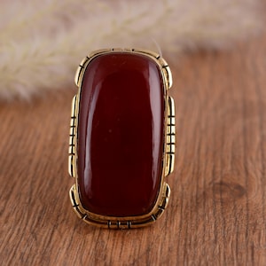 May include: A rectangular ring with a large, polished, deep red stone set in a gold-toned metal frame. The frame has a decorative, geometric design. The ring is displayed on a wooden surface, highlighting its rich colour and design.