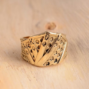 May include: A gold-coloured ring with a playing card design, showing the Ace, King, Queen, and Jack of Spades. The ring has a detailed texture and is set against a light wooden surface.