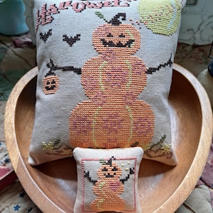 May include: Two decorative, cross-stitched pillows in a wooden bowl. The larger pillow features a pumpkin snowman design with the word "Halloween" and a bat. The smaller pillow has a similar pumpkin snowman design.