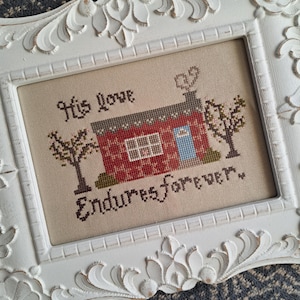 Peut inclure: Point de croix encadré représentant une maison rouge avec un toit marron et une porte bleue, avec le texte "His love Endures forever." Le cadre blanc présente des motifs floraux décoratifs. Le fond est un tissu gris texturé.