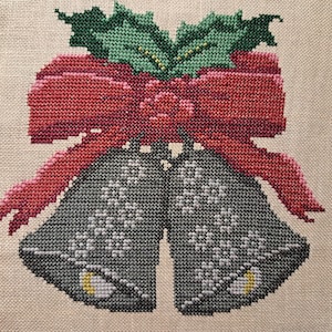 May include: Cross-stitch design of two gray bells with white snowflake patterns, a red bow, and green holly leaves. The bells have yellow accents and are set against a beige background.