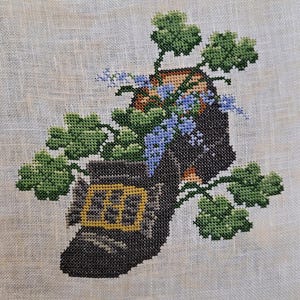 May include: Cross-stitch embroidery of a black boot with a yellow window, filled with blue flowers and green leaves. The boot is set against a neutral linen background, showcasing the detailed needlework.