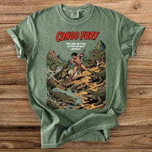 May include: Green t-shirt featuring a vintage comic book illustration. The graphic depicts a muscular figure fighting crocodiles in a jungle setting, with the text "CONGO FURY" and "SAVAGE ACTION IN THE DEADLY JUNGLE".