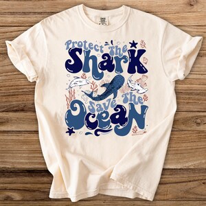 Op de afbeelding: Crèmekleurig T-shirt met de tekst "Protect the Shark Save the Ocean" in blauw. Het ontwerp bevat illustraties van haaien, koraal en andere oceaan elementen. Een comfortabel, casual T-shirt.