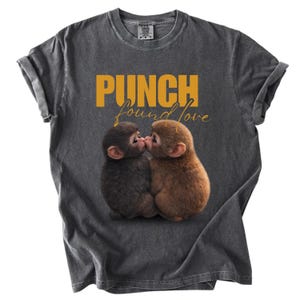 以下が含まれることがあります： ゴールドの文字で「PUNCH found love」と書かれた濃いグレーのTシャツ。2匹のキスをする赤ちゃん猿のグラフィックが特徴です。Tシャツは袖をまくり、クルーネックになっています。
