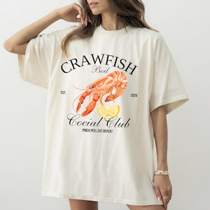 Puede incluir: Camiseta color crema con un diseño de "Crawfish Boil". El gráfico incluye una ilustración detallada de un cangrejo de río, una rodaja de limón y las palabras "Crawfish Boil Social Club" con el lema "Pinch.Peel.Eat.Repeat."