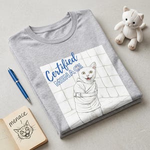 May include: A heather gray t-shirt featuring a graphic of a white cat wearing a straightjacket against a padded wall background. The text "Certified MENACE" is printed in blue above the cat. A blue pen and a small white cat plush toy are nearby.
