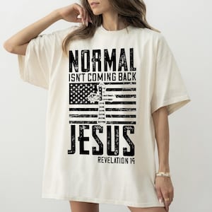 Può includere: T-shirt color crema con grafica invecchiata. Il design presenta le parole "NORMAL ISN'T COMING BACK" sopra una bandiera americana stilizzata con una croce, e la parola "JESUS" sotto, con "REVELATION 14" in basso.