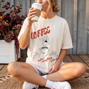 May include: A cream-coloured short-sleeve t-shirt featuring a graphic print with the red text COFFEE and black line art of a hand pouring liquid into a cup, a croissant, and cookies, with the red text makes everything better below the cup.