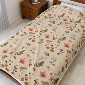 May include: A beige blanket with a repeating pattern of mushrooms, leaves, and berries in shades of brown, red, and gray. The blanket has a white border and is placed on a bed. The overall theme is autumn and nature.