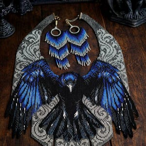 Puede incluir: Un conjunto de collar y pendientes de cuentas. El collar presenta un diseño de cuervo negro con alas azules, sobre un fondo gris y plateado. Los pendientes tienen flecos azules y plateados.