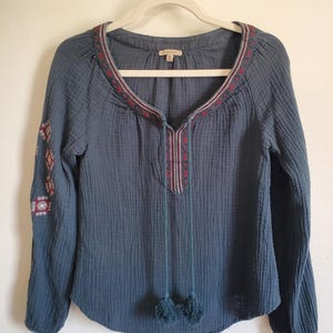 May include: A blue long-sleeve blouse with embroidered details on the sleeves and neckline. The neckline features a V-neck with tassels. The blouse is hanging on a white hanger against a white wall.