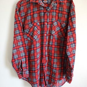 May include: Red, blue, and gray plaid button-down shirt. The shirt has two chest pockets and a collar. The Dickies brand label is visible on the collar. The shirt is hanging on a blue hanger.