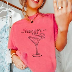 May include: A coral-colored t-shirt featuring a black graphic of a martini glass with a lemon slice and the text "FIRSTWIVES Club ESTD 1995". The shirt has short sleeves and a crew neck.
