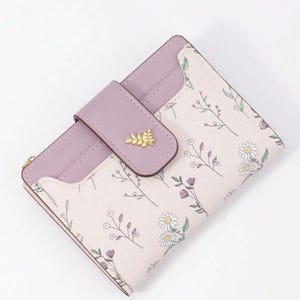 May include: A small, bi-fold wallet with a floral pattern. The wallet features a light pink background with delicate floral sprigs in white, purple, and green. It has a lavender flap with a gold leaf accent and a snap closure. The wallet is approximately 4 inches wide.
