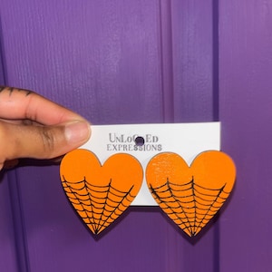 Pode incluir: Um par de brincos em forma de coração laranja com um design de teia de aranha preta. Os brincos estão presos a um cartão de exibição branco com o texto UNLOCKED EXPRESSIONS impresso em preto, incluindo um pequeno ícone de aranha.